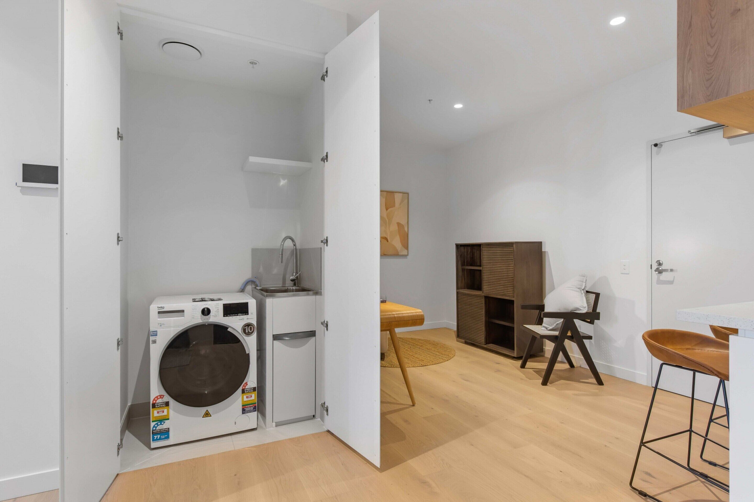 A compact living space with a built-in washer and dryer, a small kitchenette, a wooden floor, and a seating area with a chair and stool.