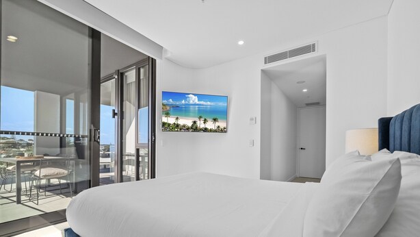 A hotel room with a large bed, a balcony with a glass door, a mounted TV, and a view of the beach. - Stylish 2-bed Apartment With Rooftop BBQ Access (Perth)