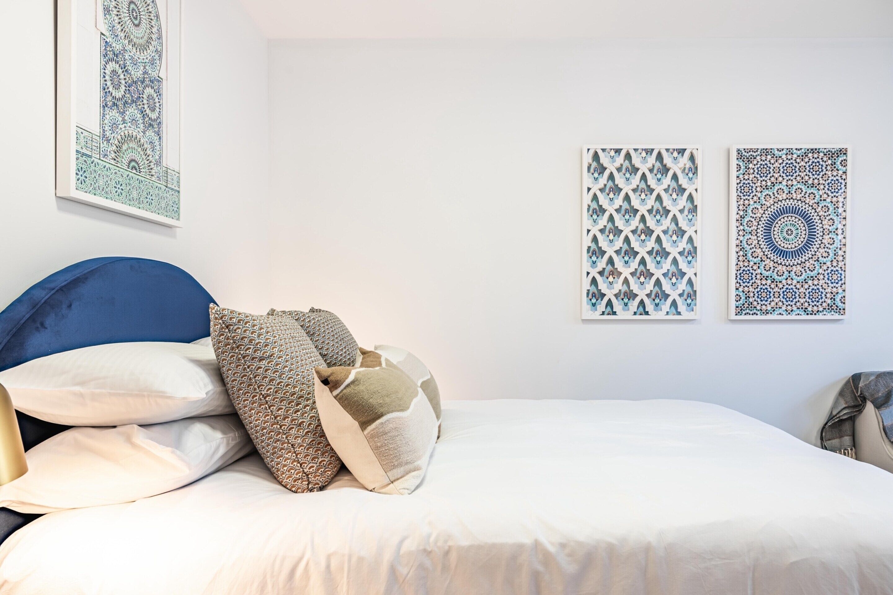Un lit bien fait, avec une tête de lit bleue, du linge de lit blanc et des coussins à motifs. Deux œuvres d’art encadrées sont accrochées au mur.
