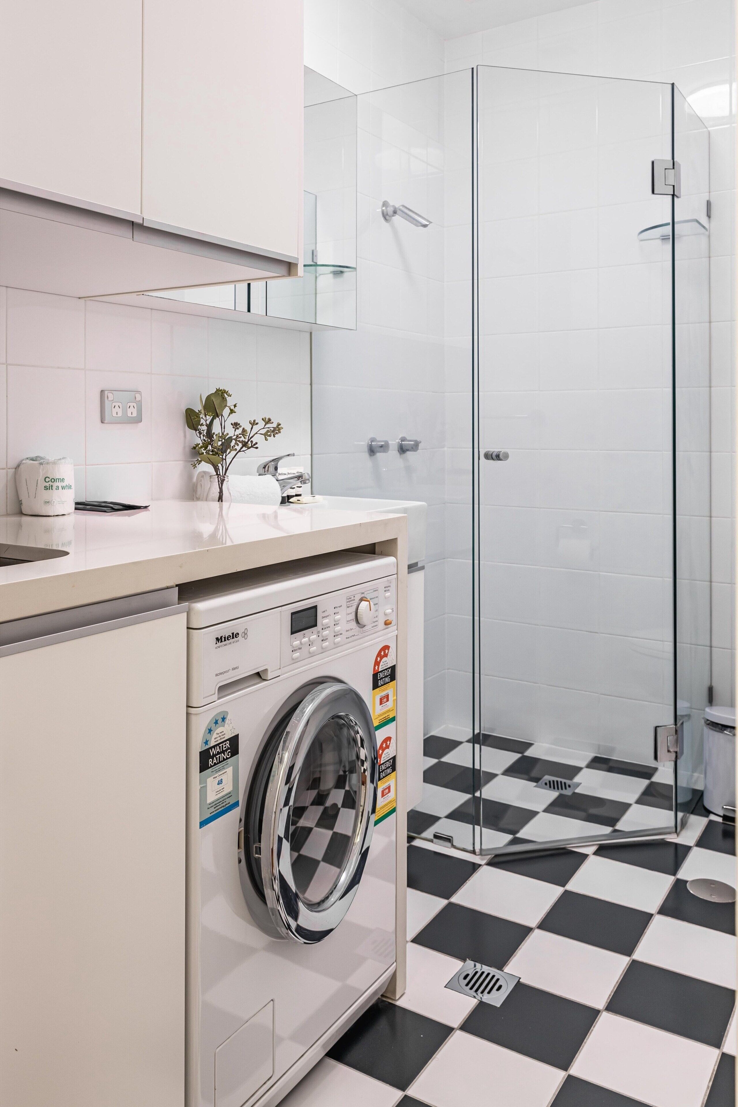 Une salle de bain moderne équipée d’un lave-linge, d’une douche à parois de verre et d’un carrelage à motifs carrés.