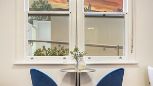 Una habitación con dos sillas azules, una mesita redonda con un florero y una ventana con vista a un edificio y árboles.