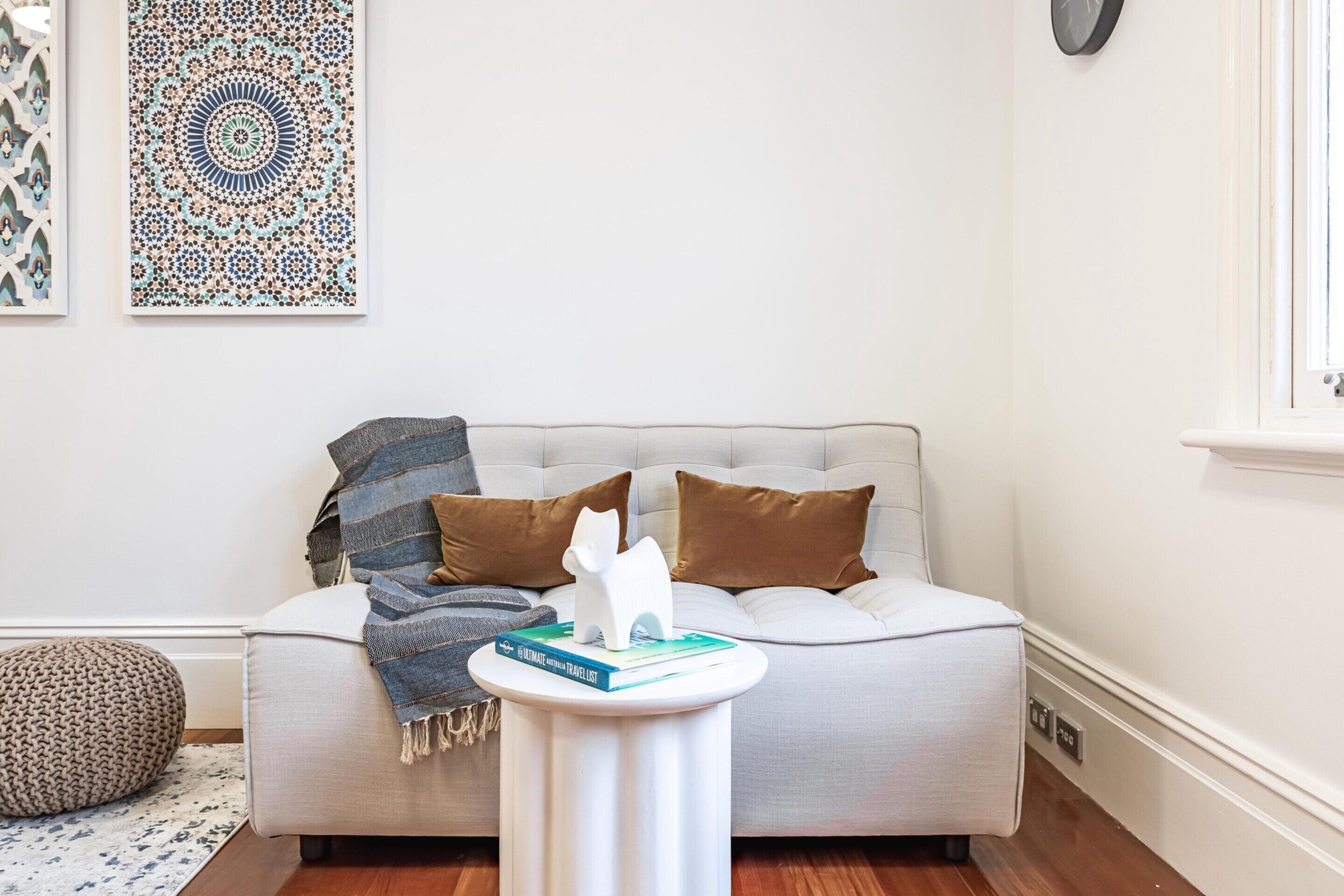 A living room with a sofa, a round white side table, a patterned wall hanging, and a window with a clock.