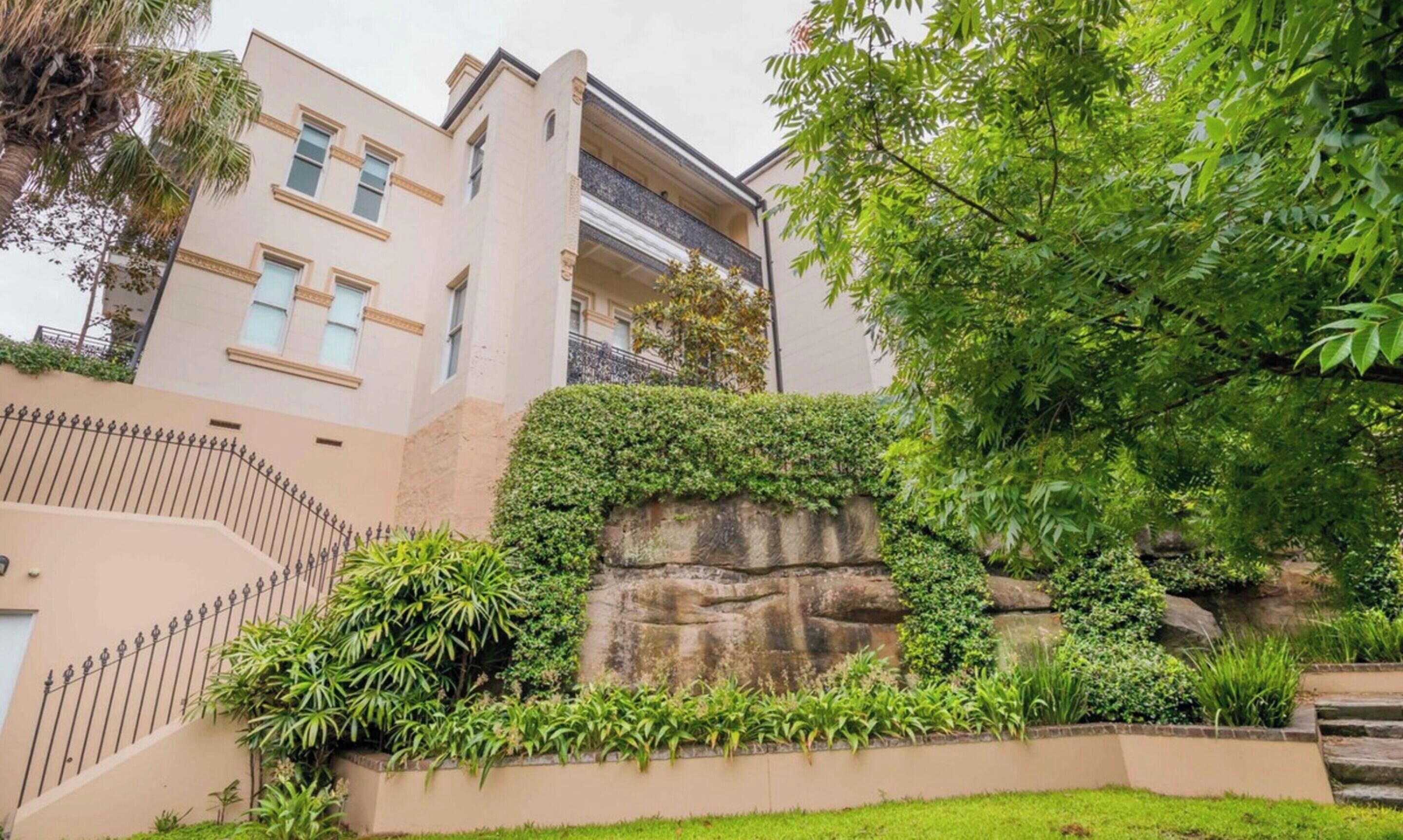 Un edificio de varios pisos con fachada color beige, un muro de piedra de contención y diversa vegetación, incluyendo un árbol grande y varios arbustos.