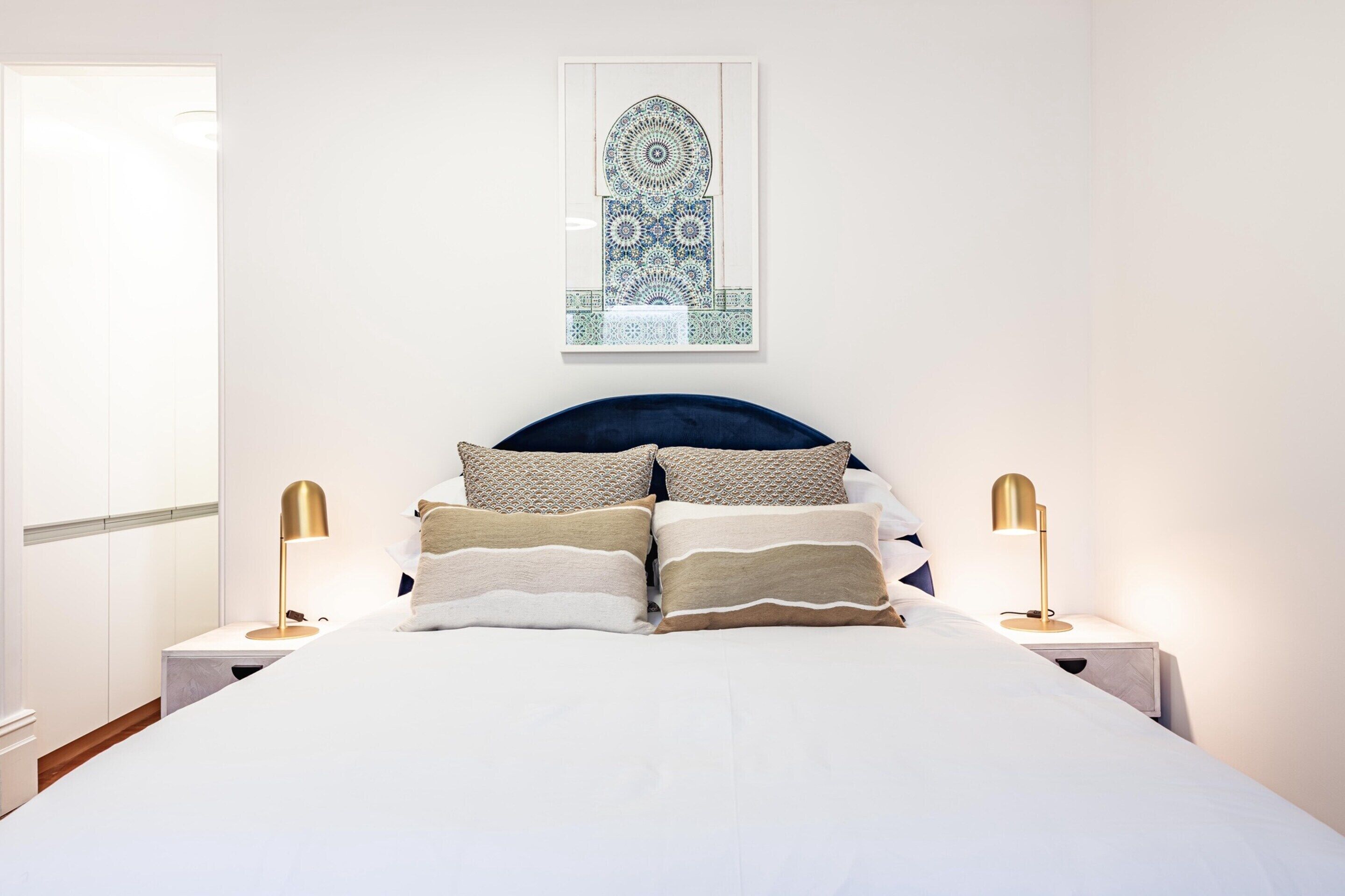 A neatly made bed with a dark headboard, two bedside lamps, and a framed artwork above the bed.