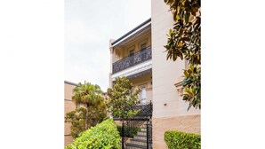 Un edificio con un camino a cuadros que lleva a una entrada con portón, rodeado de follaje y una palmera.