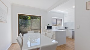 A modern kitchen with white cabinetry, a central island, and a dining area with a glass table. - Elegant 3-bed Home From Sunny Glenelg Beach (Adelaide)