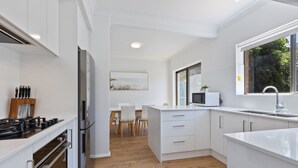 A modern kitchen with white cabinetry, a built-in oven, a microwave, and a large window offering a view of greenery outside. - Elegant 3-bed Home From Sunny Glenelg Beach (Adelaide)