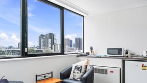 Una habitación moderna con un ventanal grande, un sillón de cuero negro, una mesita de madera pequeña, un refrigerador blanco y un microondas sobre una encimera.