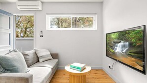 A modern living room with a sofa, a round wooden coffee table, a flat-screen TV, and a window with a view of trees.