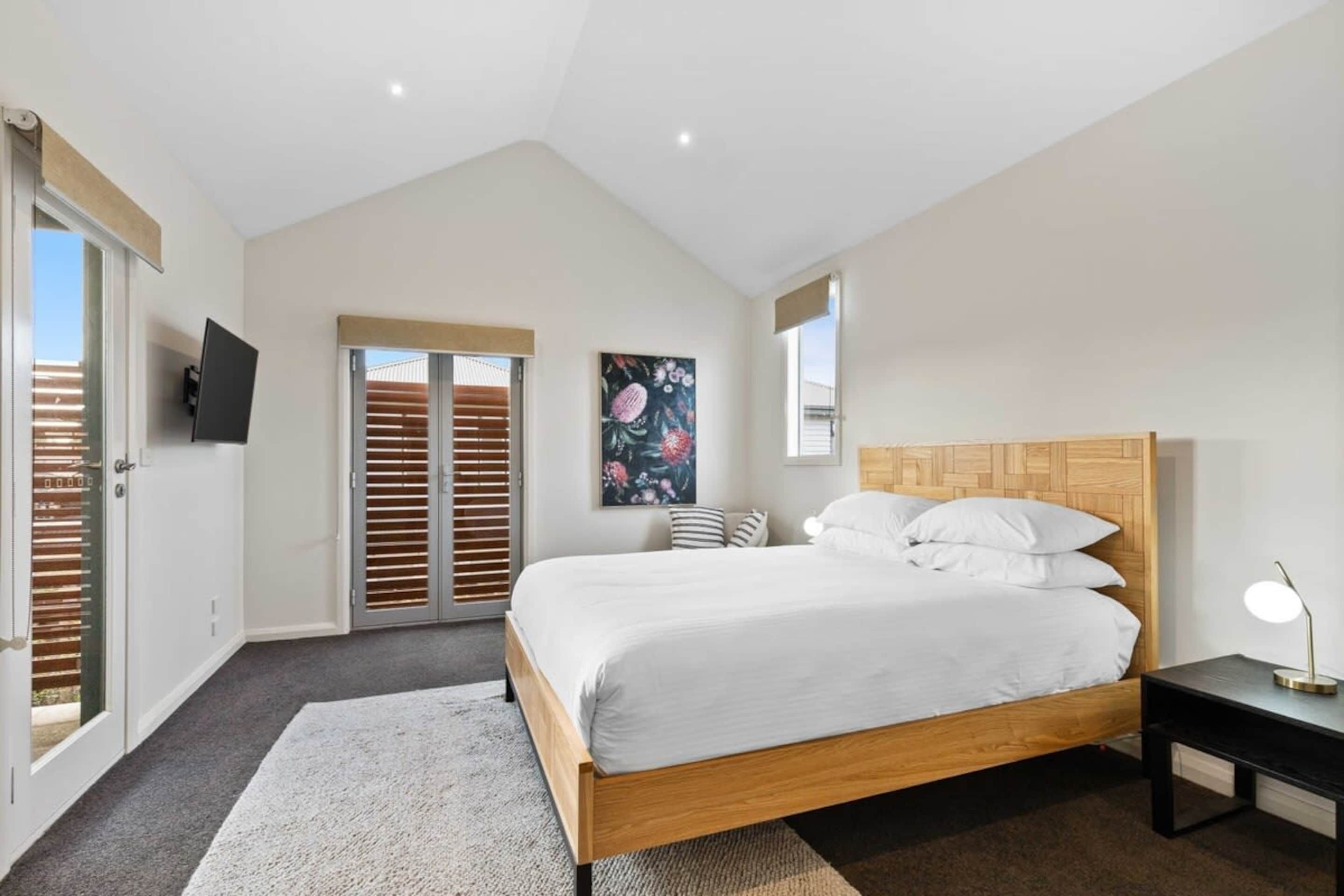 A bedroom with a large bed, a desk, a TV mounted on the wall, and a window with blinds.