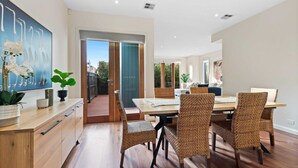 A modern dining area with a wooden table and wicker chairs, a large painting on the wall, and a view through a glass door. - Chic 2-bed Home By Bustling Shops (Melbourne)