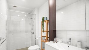 A modern bathroom with a glass-enclosed shower, a white toilet, a wooden shelf with a plant, and a white sink with a mirror. - Modern 2-bedroom Retreat Minutes From St Kilda (Melbourne)
