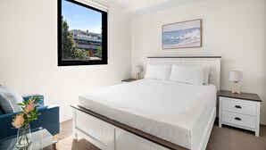 A bedroom with a large bed, a nightstand with two lamps, a window with a view of buildings, and a painting on the wall. - Modern 2-bedroom Retreat Minutes From St Kilda (Melbourne)