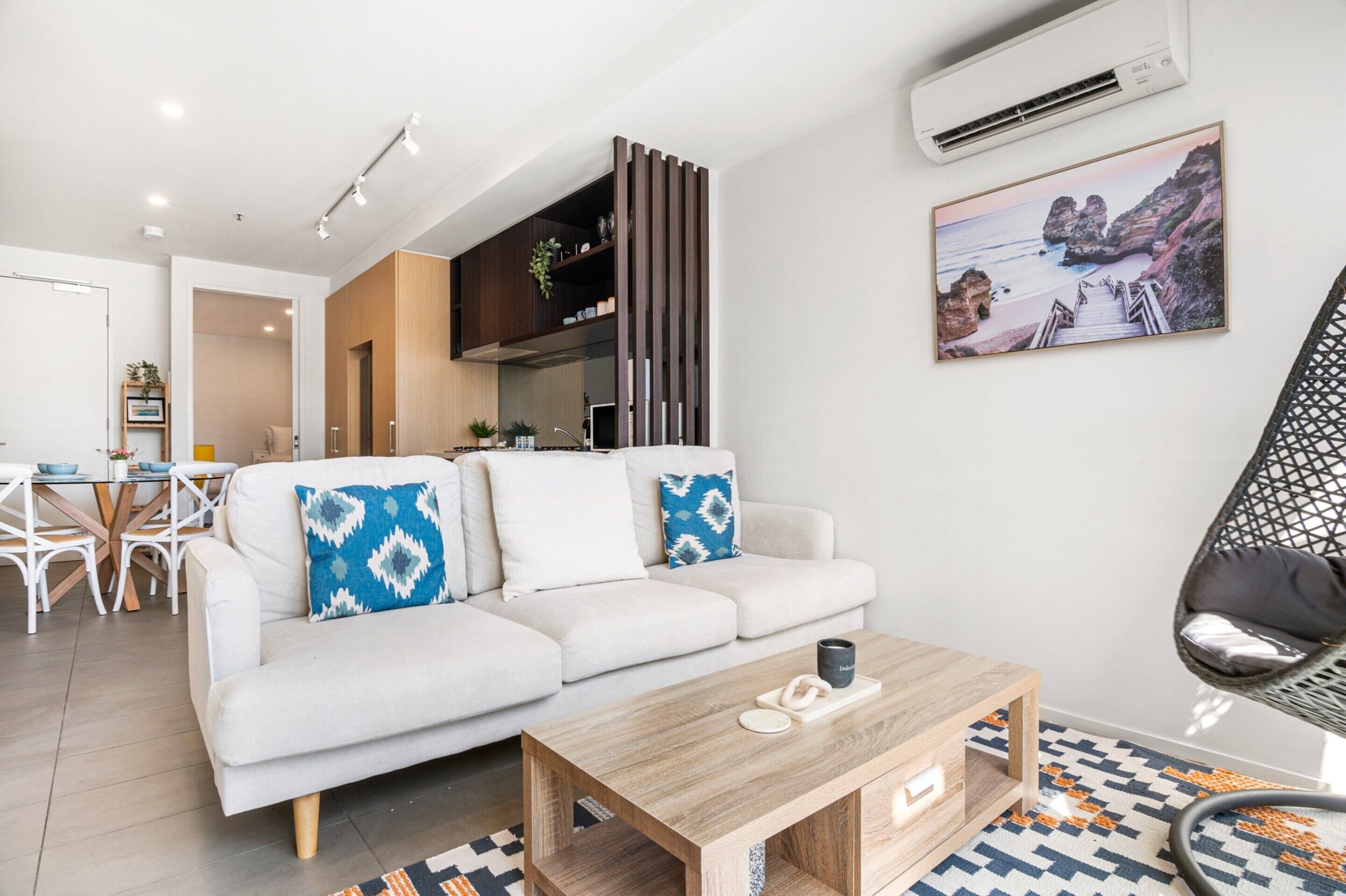 A modern living room with a sofa, coffee table, and a hanging chair. There is a dining area with a table and chairs, and an air conditioning unit on the wall.