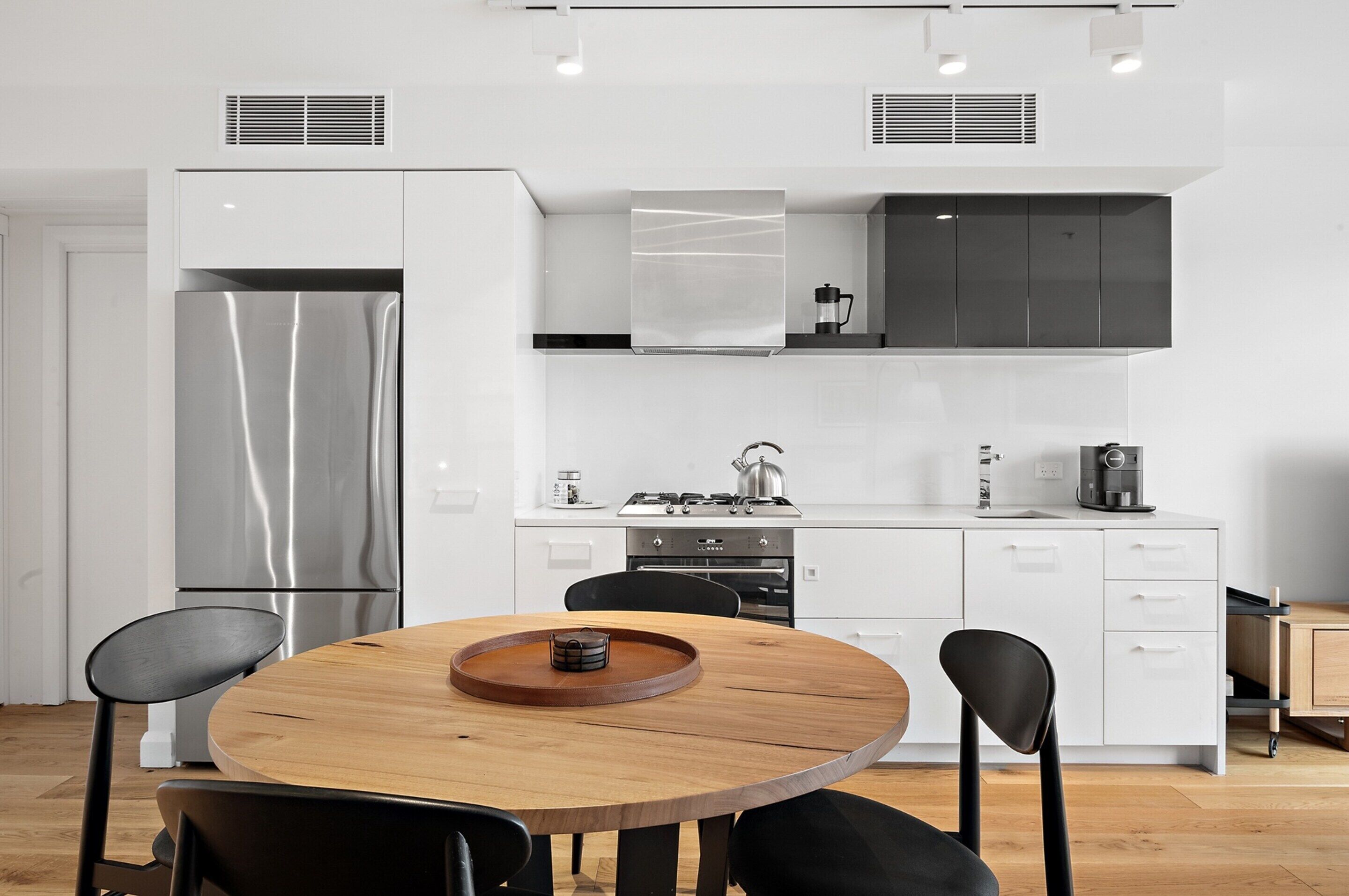 A modern kitchen with white cabinetry, a stainless steel refrigerator, a gas stove, and a wooden dining table with chairs.