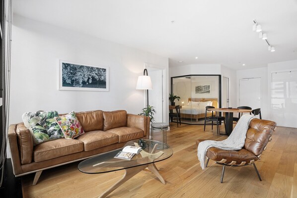 A modern living room with a leather sofa, glass coffee table, and a leather armchair. There is a dining area with a table and chairs in the background.