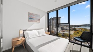 A modern hotel room with a large bed, a nightstand with a lamp, a chair, and a large window offering a view of buildings and a body of water. - Sleek 2-bed Apartment With Rooftop Garden Access (Canberra)