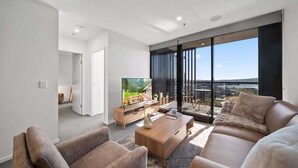 A modern living room with a sofa, armchairs, a coffee table, and a TV. There is a large window with a view of a cityscape and a balcony. - Sleek 2-bed Apartment With Rooftop Garden Access (Canberra)