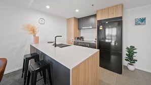A modern kitchen with a central island, black stools, a clock, and a potted plant. - Sleek 2-bed Apartment With Rooftop Garden Access (Canberra)