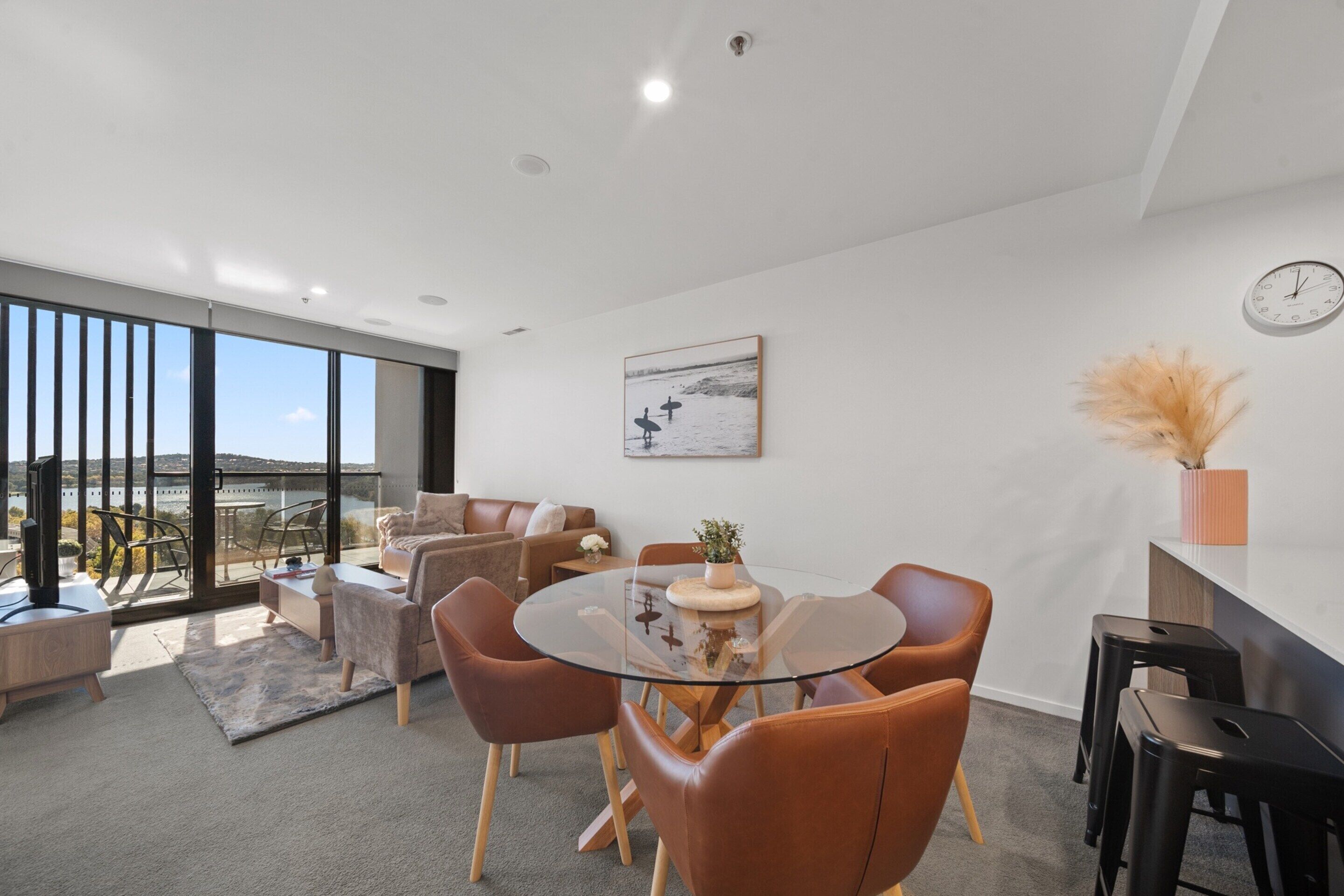 A modern living room with a glass dining table surrounded by leather chairs, a sofa, and a coffee table. There is a large window offering a view of the outdoors.
