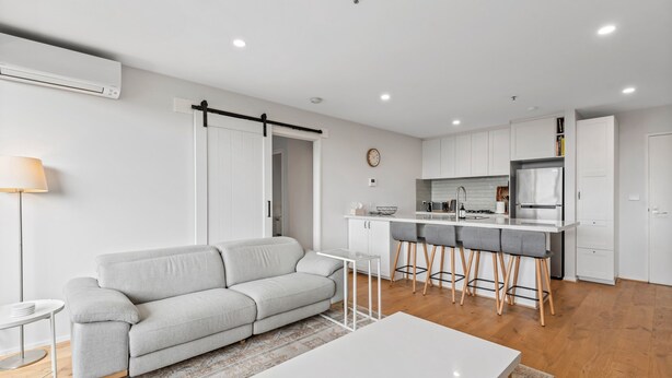 A modern living room with a grey sofa, a white coffee table, and a kitchen area with a bar counter and stools. - Sleek Kensington 2-bed With BBQ Area & Gym (Melbourne)