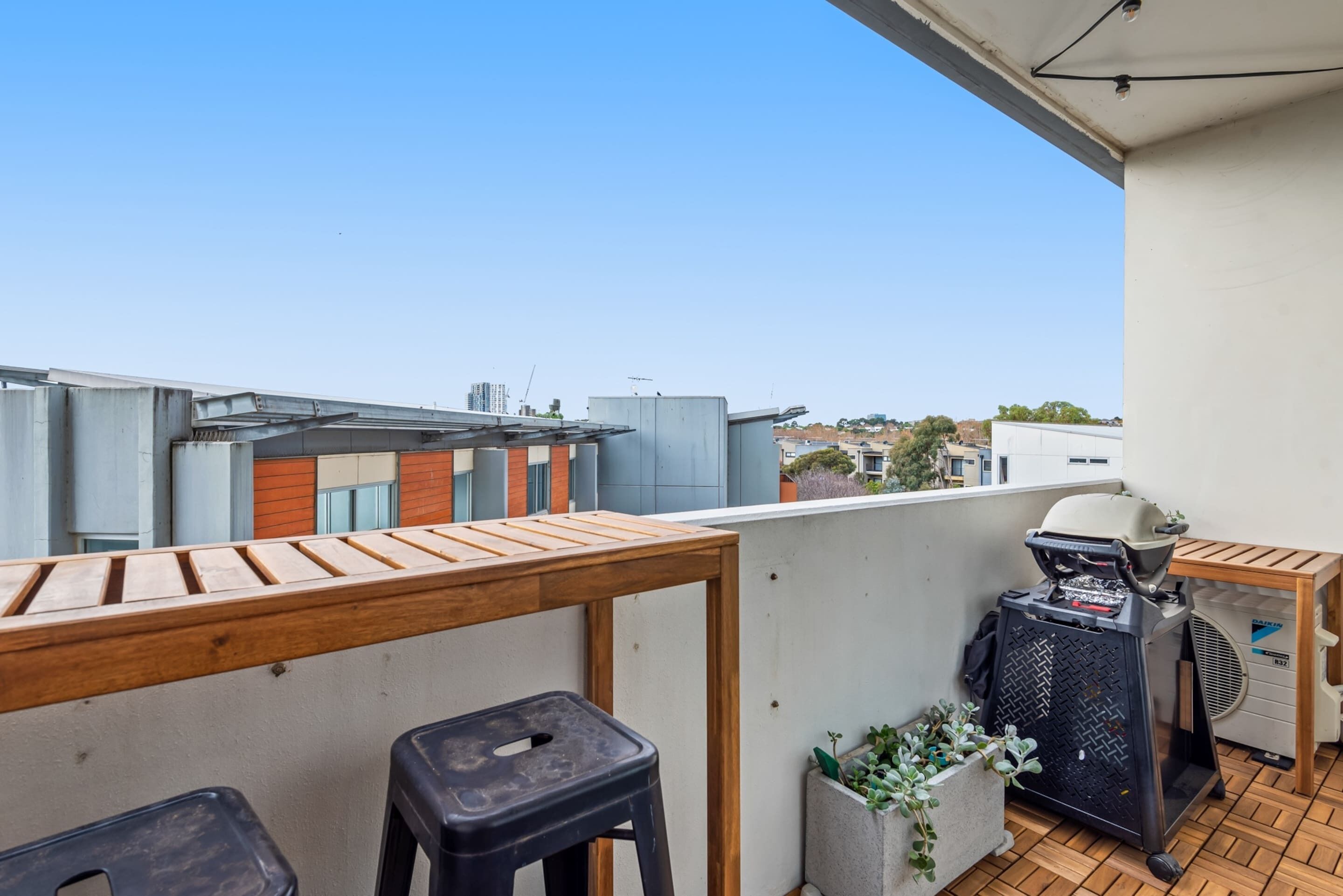 Ein Balkon mit einem Holztisch, zwei schwarzen Hocker, einem Grill und einem Blumentopf.