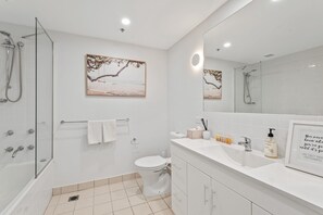 A modern bathroom with a glass shower enclosure, a toilet, a sink with toiletries, and two wall-mounted paintings. - Sleek Kensington 2-bed With BBQ Area & Gym (Melbourne)