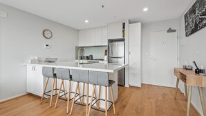 A modern kitchen with a central island, bar stools, a refrigerator, and a dining area with a table. - Sleek Kensington 2-bed With BBQ Area & Gym (Melbourne)