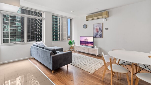 Ein modernes Wohnzimmer mit grauem Sofa, Fernseher, Esstisch mit Stühlen und großen Fenstern mit Jalousien.