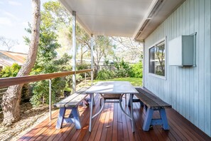 Une terrasse couverte avec une table de pique-nique et des bancs, entourée d’arbres et d’une maison.