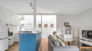 A modern kitchen with white cabinets, a blue island, and a built-in oven. There is a grey sofa and a TV in the living area. - Cosy Getaway With Fireplace and Spacious Backyard (Broulee)
