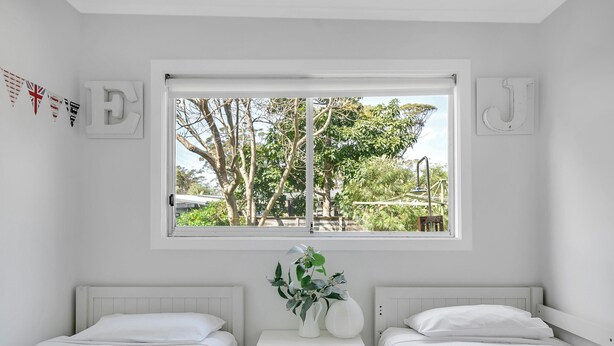 A bedroom with two beds, a window with a view of trees and a park, and a small table with a vase and a plant. - Cosy Getaway With Fireplace and Spacious Backyard (Broulee)