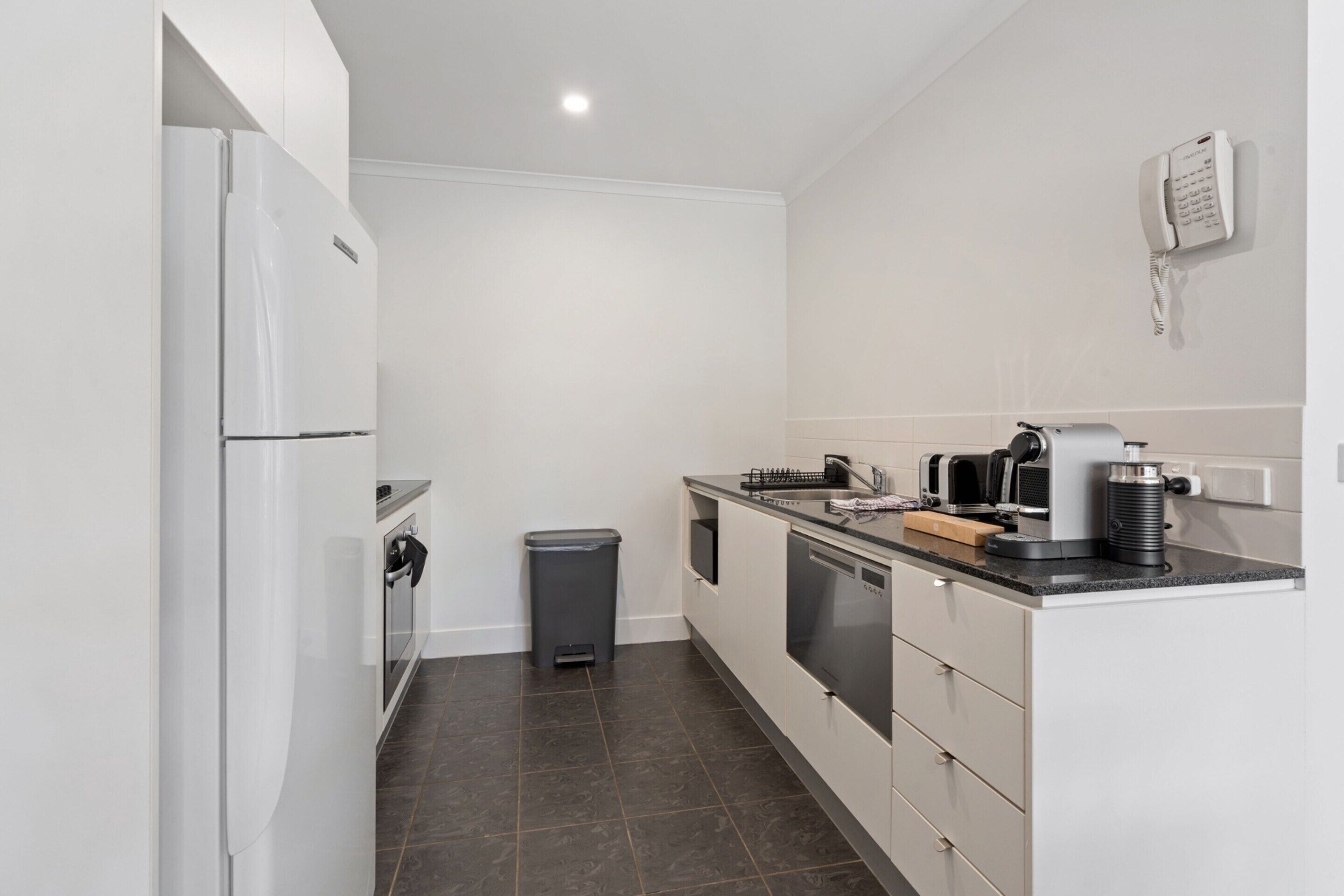 A modern kitchen with white cabinets, a black countertop, a coffee machine, and a trash bin.