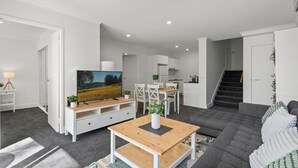 A modern living room with a flat-screen TV, a wooden coffee table, a grey sofa, and a dining area with white chairs and a small kitchen in the background. - Central Braddon Retreat With Balcony & Pool Access (Canberra)