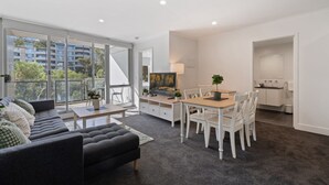 A modern living room with a sofa, dining table, and chairs. There is a television and a painting on the wall. The room has large windows and a view of buildings outside. - Central Braddon Retreat With Balcony & Pool Access (Canberra)