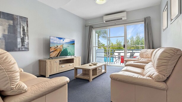 Un salon moderne avec des canapés de couleur beige, une table basse en bois et un téléviseur à écran plat posé sur un meuble en bois. De grandes fenêtres offrent une vue sur l’extérieur.