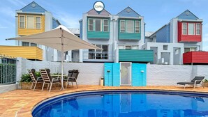 Une piscine avec une terrasse en briques, entourée d’un mur blanc et d’une clôture. On y trouve des chaises longues et un espace ombragé.