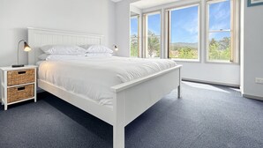Une chambre à coucher avec un lit blanc, deux tables de chevet avec des lampes, une grande fenêtre et une vue sur la verdure.