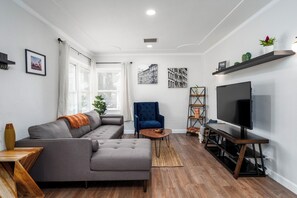 Ein modernes Wohnzimmer mit einem grauen Ecksofa, einem blauen Sessel, einem Holztisch, einem Fernseher auf einem Schrank und einem Bücherregal.