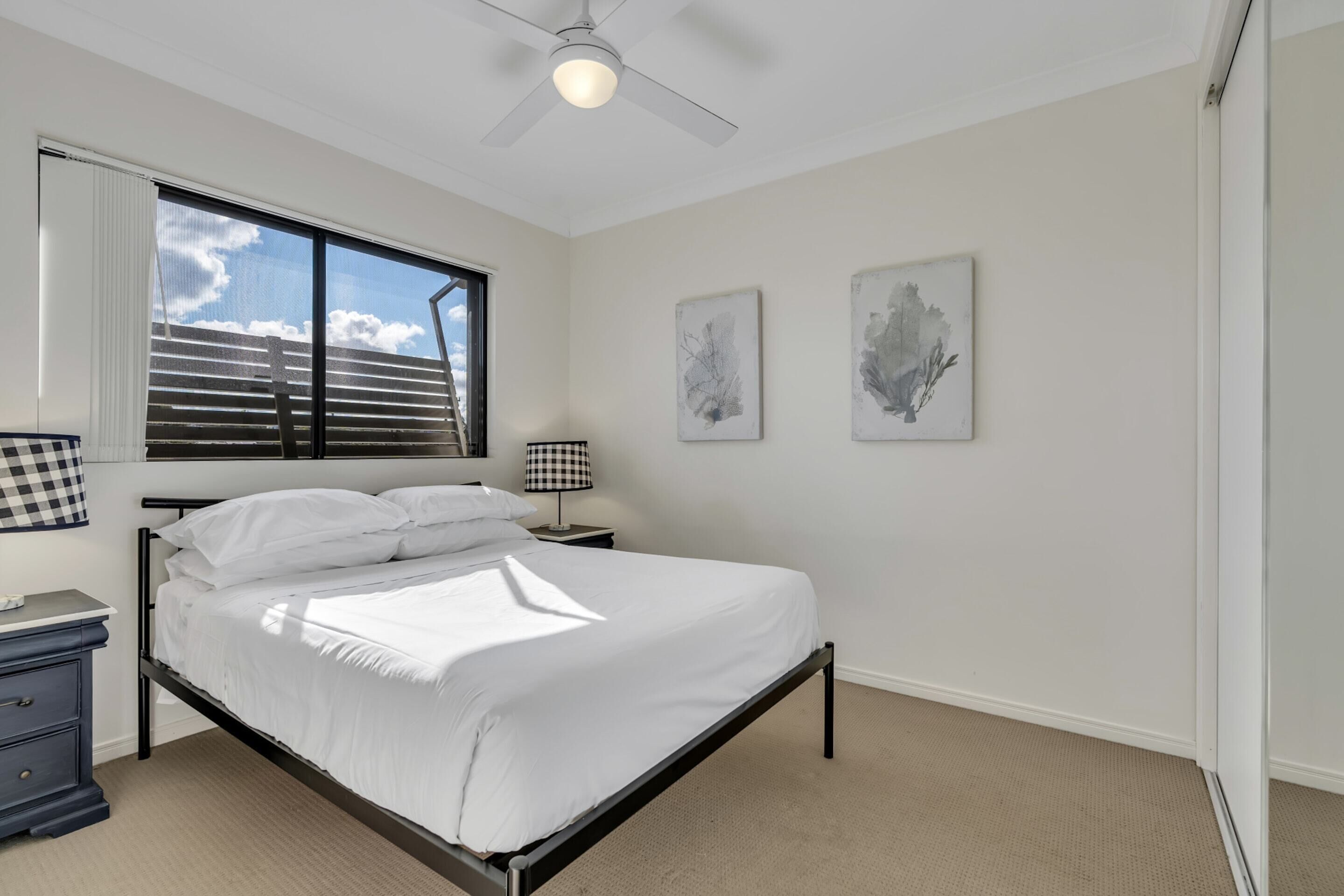 A bedroom with a bed, a ceiling fan, a window with blinds, and two wall-mounted artworks.