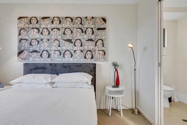 A modern bedroom with a large bed, a headboard, a wall art piece, a bedside table with a lamp, and a view into the bathroom. - Bright 2-bed With Private Balcony & Serene View (Brisbane)