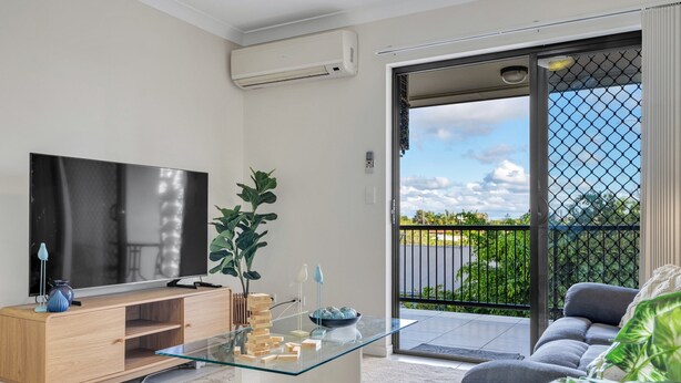A modern living room with a glass coffee table, a flat-screen TV, a wooden TV stand, and a sliding glass door leading to a balcony with a view of greenery. - Bright 2-bed With Private Balcony & Serene View (Brisbane)
