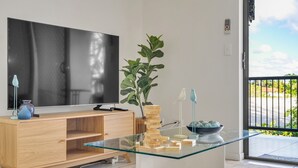 A modern living room with a glass-top coffee table, a wooden TV stand, a large flat-screen TV, a potted plant, and a view of the outdoors. - Bright 2-bed With Private Balcony & Serene View (Brisbane)