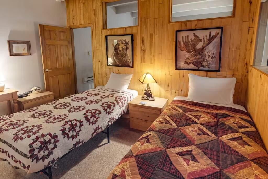 A room with two beds, wooden walls, a small bedside table with a lamp, and framed pictures on the wall.