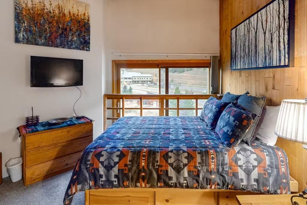 A bedroom with a bed, a dresser, a TV, and a window with a view of a building and landscape.