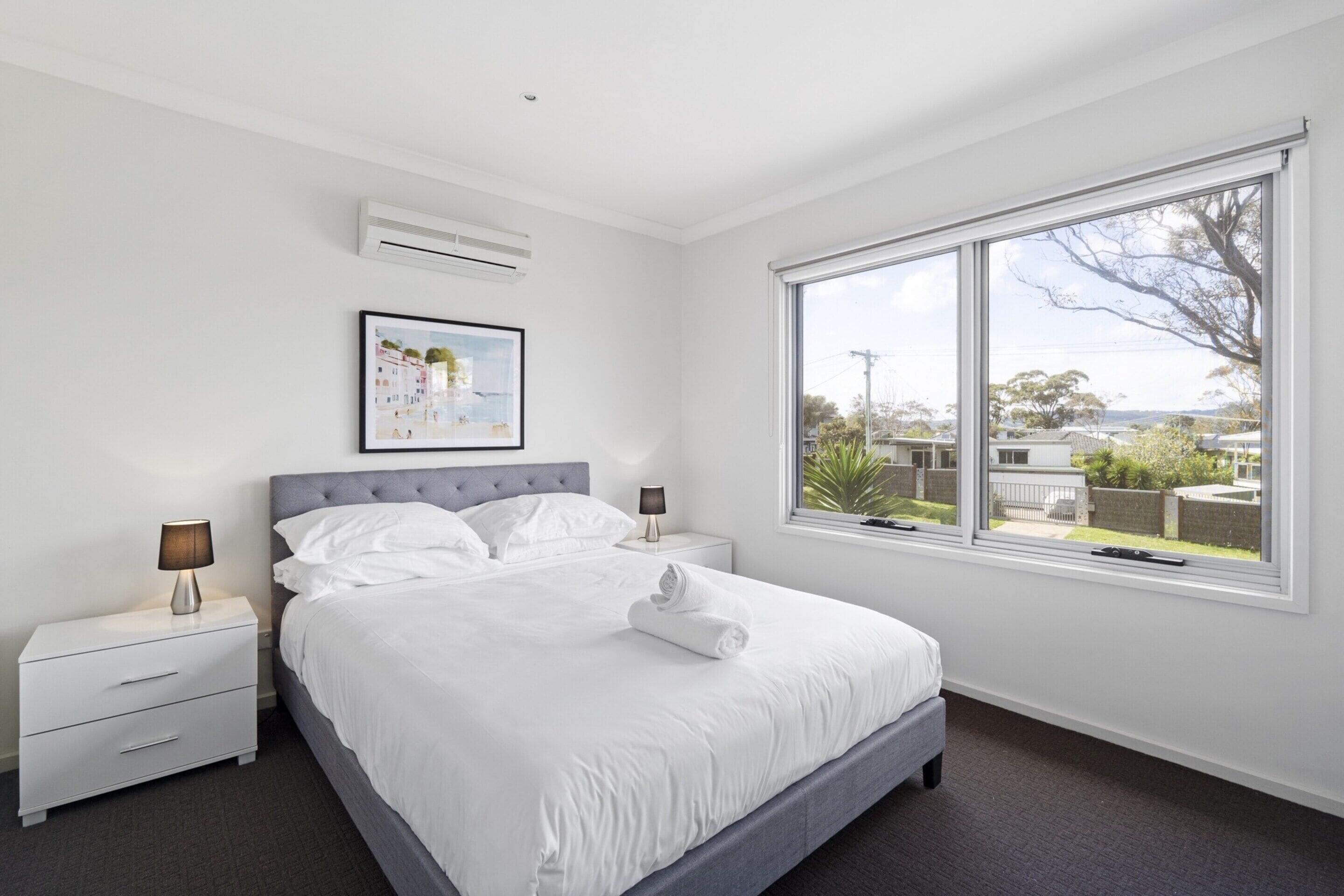 A modern bedroom with a large bed, two bedside tables, a framed picture, and a large window with a view of the outdoors.