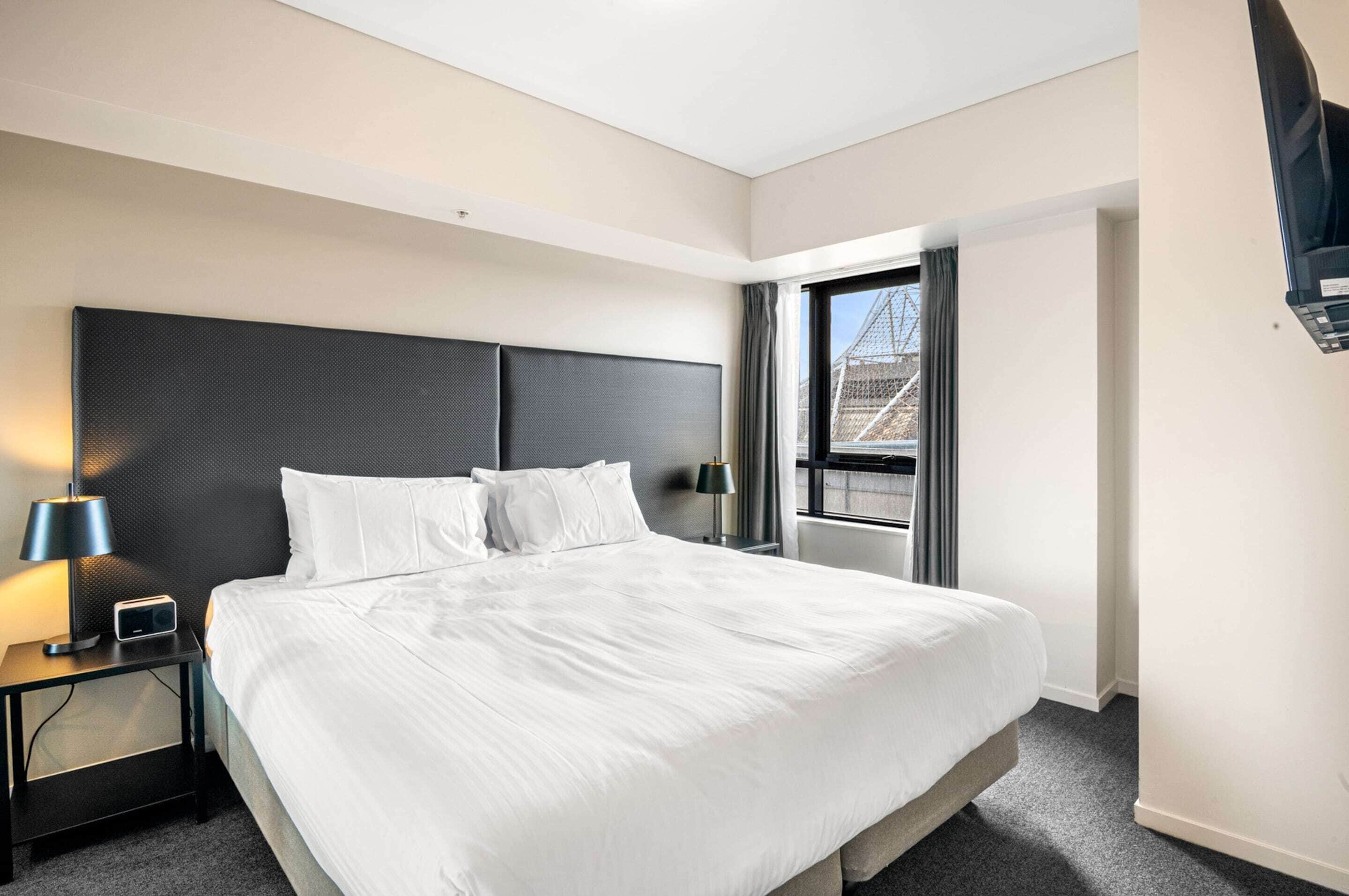A modern hotel room with a large bed, a nightstand with a lamp, and a window with curtains.
