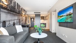 A modern living room with a grey sofa, a round white table with a plant, a large TV on the wall, and a kitchen in the background. - Spacious Apartment in Heart of Southbank (Melbourne)