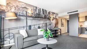 A modern hotel room with a grey sofa, a round table with a potted plant, a cityscape mural on the wall, and a kitchenette area. - Spacious Apartment in Heart of Southbank (Melbourne)
