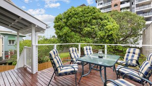 Terraza exterior con una mesa de vidrio y sillas a rayas, rodeada por una barandilla blanca y un árbol grande.